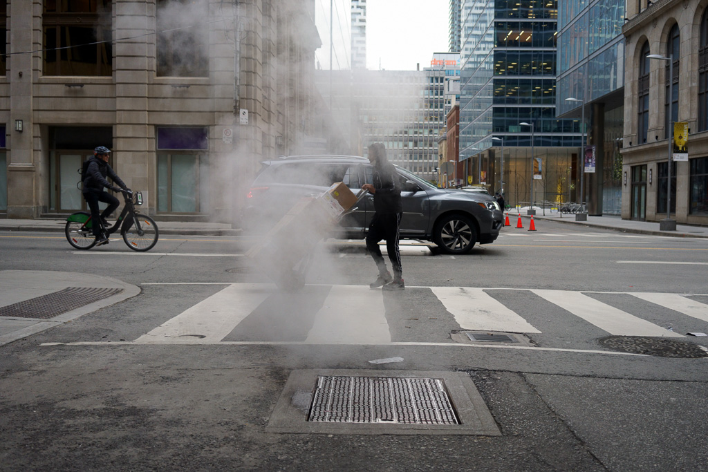 Man pushes a hand truck loaded with boxes through a blast of steam from a vent in the road.