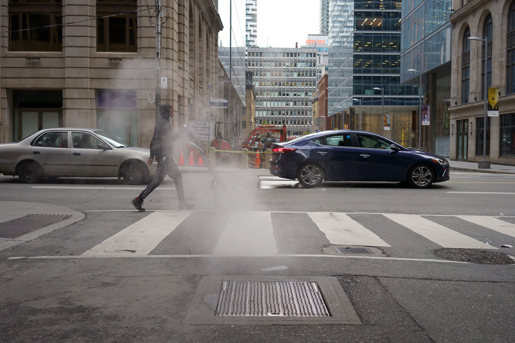 Man pushes an empty hand truck through a blast of steam from a vent in the road.