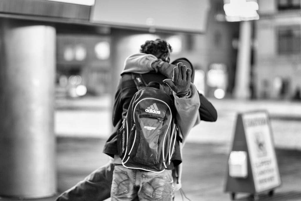 Black and white photograph of man with hand up to block the shot while he hugs another man