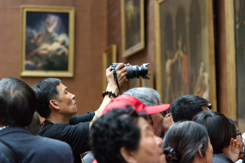 A man raises a camera over his head and photographs a painting in the Louvre museum.