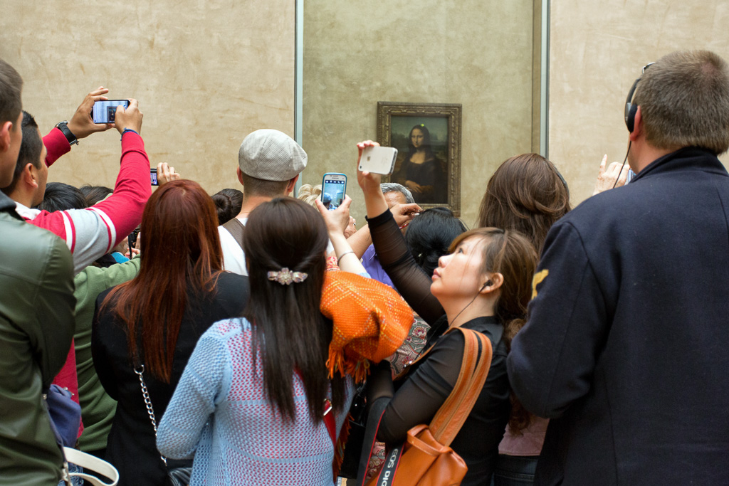 People with iPhones crowd around the Mona Lisa painting in the Louvre museum trying to take selfies with the famous portrait.