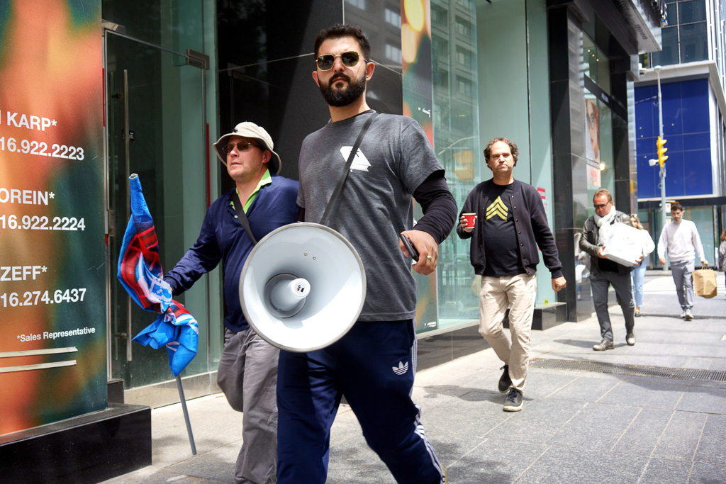 A man with a bullhorn walks down the sidewalk with a man carrying a flag.