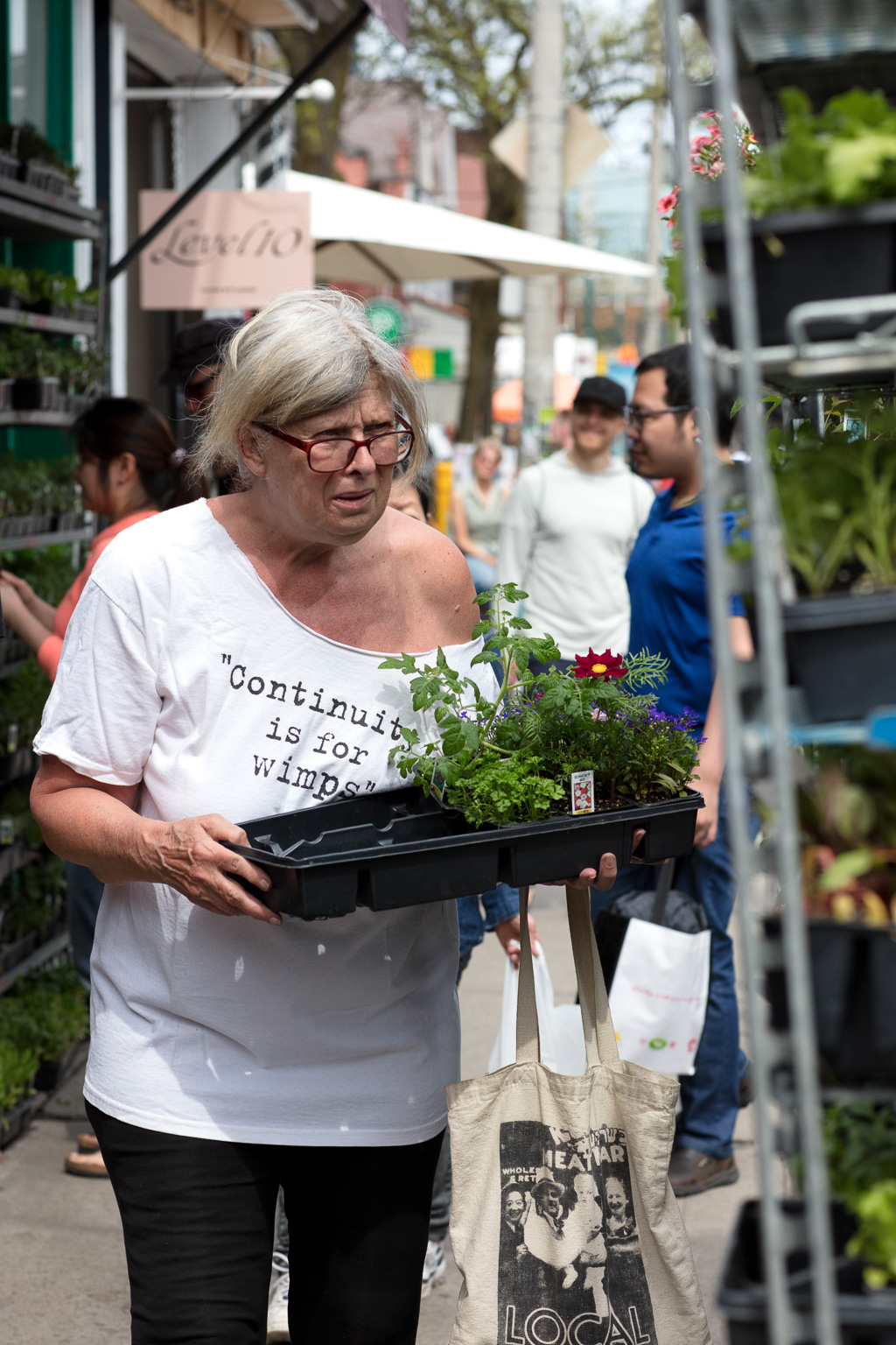 A middle-aged woman with glasses carries a plastic tray with some plants and wears a white T-shirt with the words: "Continuity is for wimps"