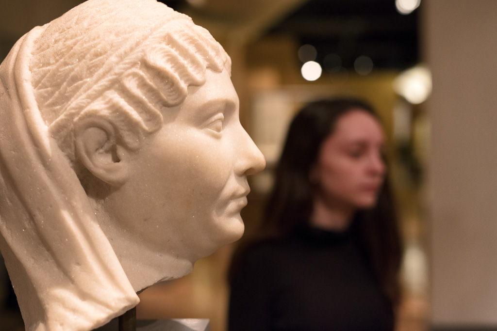 A marble bust sits in a museum gallery while a young woman passes in the background.