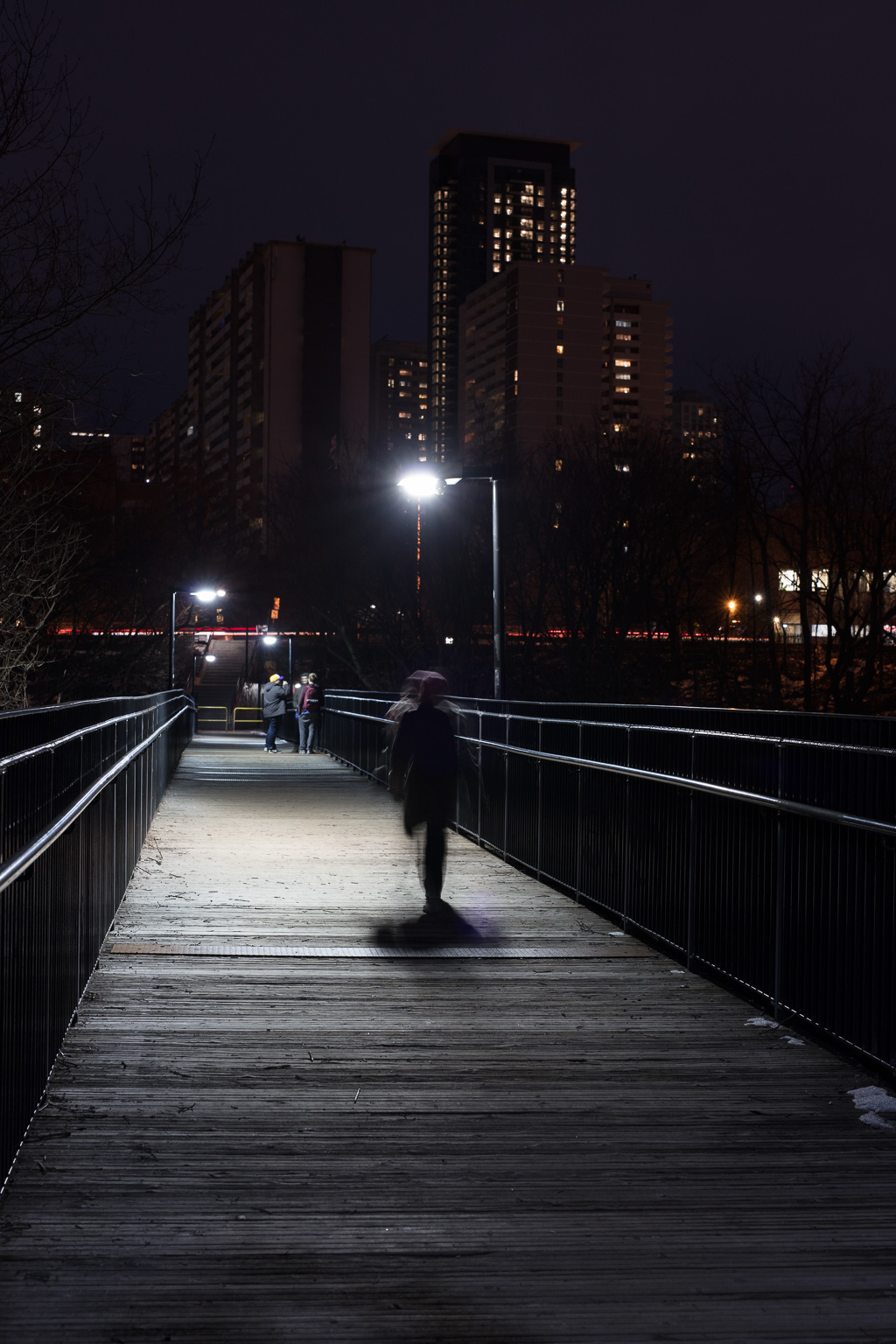 Night shot standing on Toronto's Glen Road Pedestrian Bridge. A blurred person runs towards a group of people loitering at the far end of the bridge. In the background rise the apartment buildings of St. James Town.