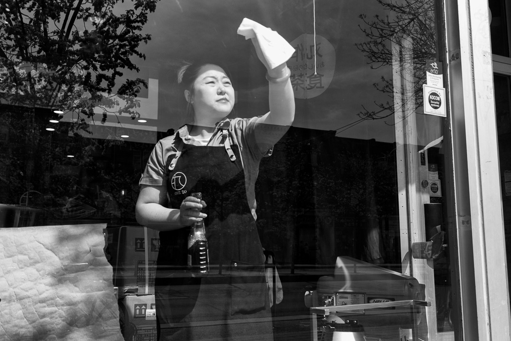 A woman cleans the interior face of a store window, holding a bottle of window cleaner in one hand and a white cloth in the other.