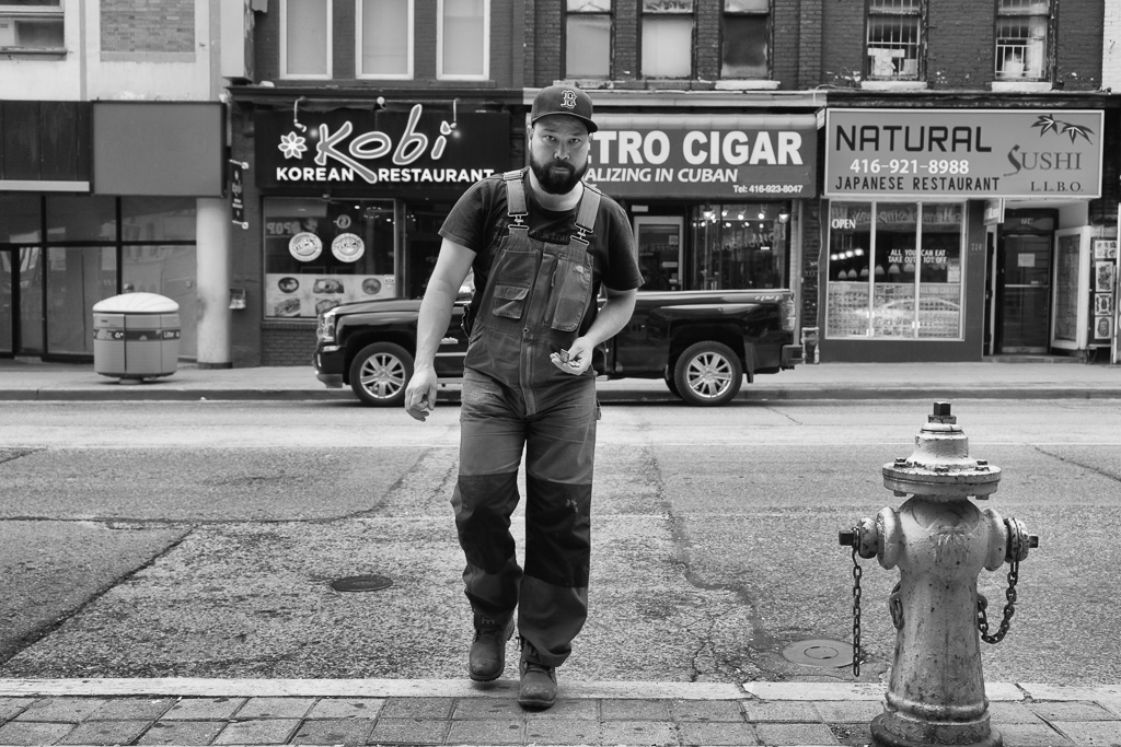 A man in overalls steps up onto the curb beside a fire hydrant while in the background are stores fronting a city street.