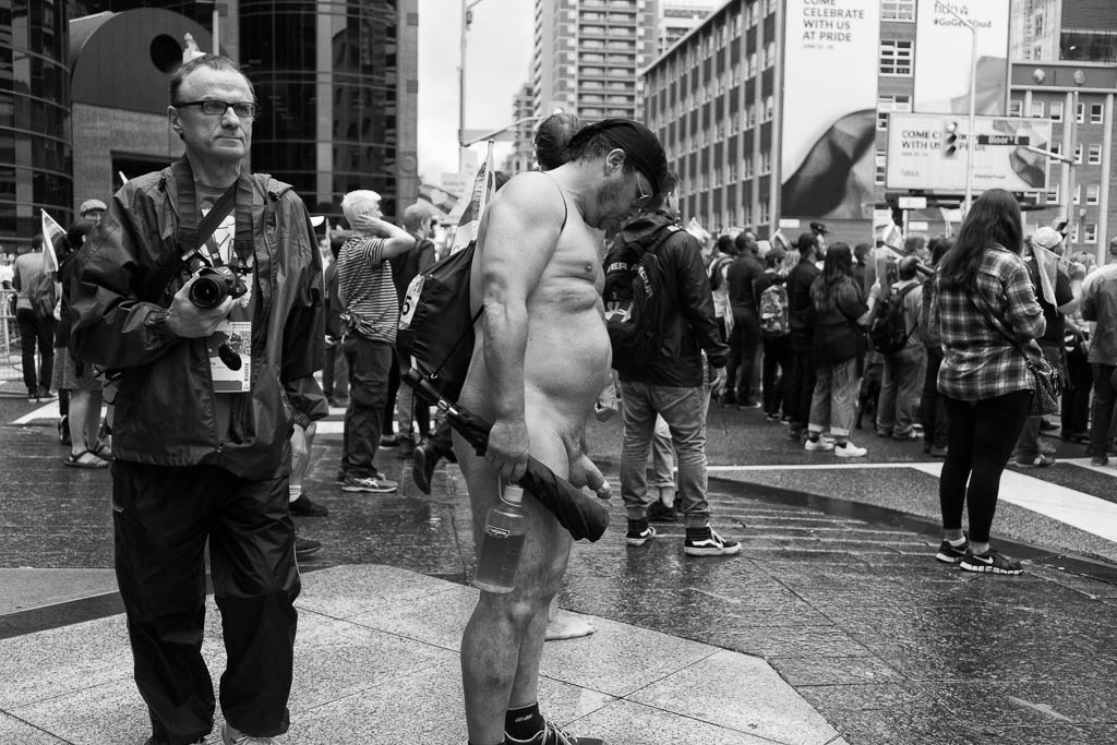 A man stands naked on a street corner and stares down at his schlong.