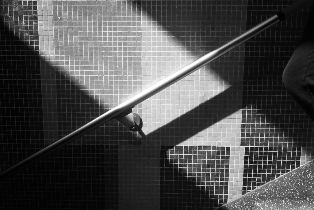 Black and white photo of a bright railing angling diagonally to the lower left corner against a backdrop of small wall tiles illuminated by a shaft of light angling diagonally to the lower right corner