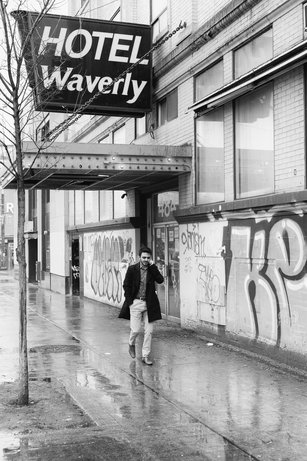 A man talking on a cell phone walks on wet pavement past the graffiti-covered entrance to the Hotel Waverly. 