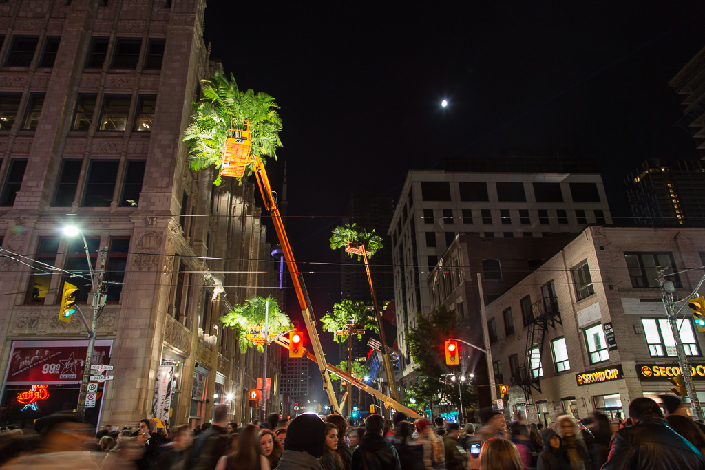 A Nuit Blanche outdoor contemporary art exhibit on Toronto's Queen Street West featuring orange cherry pickers extended high above the crowded street and laden with palm fronds.