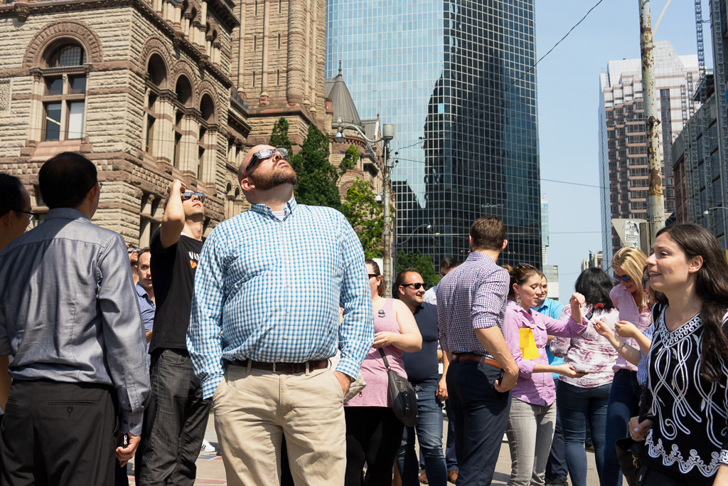 A man wearing special glasses stands in a crowd gazing at the sun. In the background is Toronto's Old City Hall on Queen Street West.