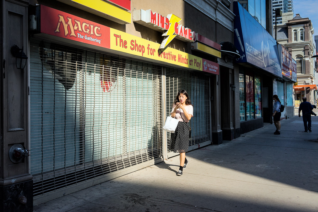 A woman walks through a sliver of morning light on Yonge Street while behind her is a shop called Mind Games with a bolt of lightning above the woman's head.