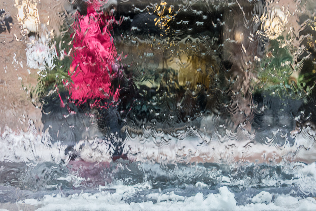 A blurred figure in a red hooded coat walks behind a glass screen as water pours down the surface of the glass.