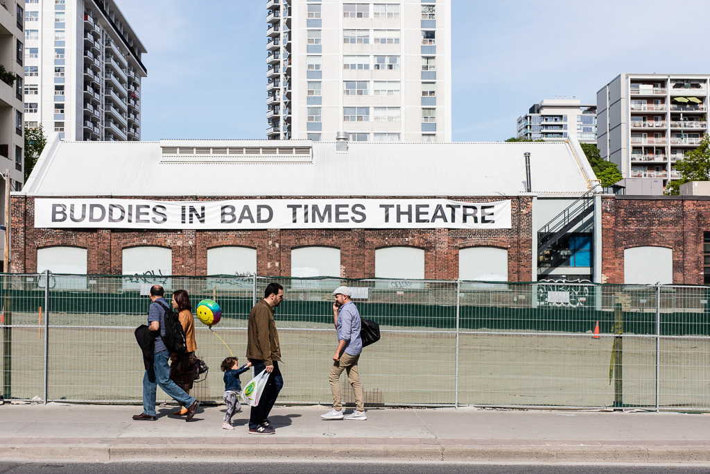 A father and small child walk on the sidewalk while holding hands. In the child's free hand is a happy face balloon. On the building in the background is a large sign that says: Buddies In Bad Times Theatre.