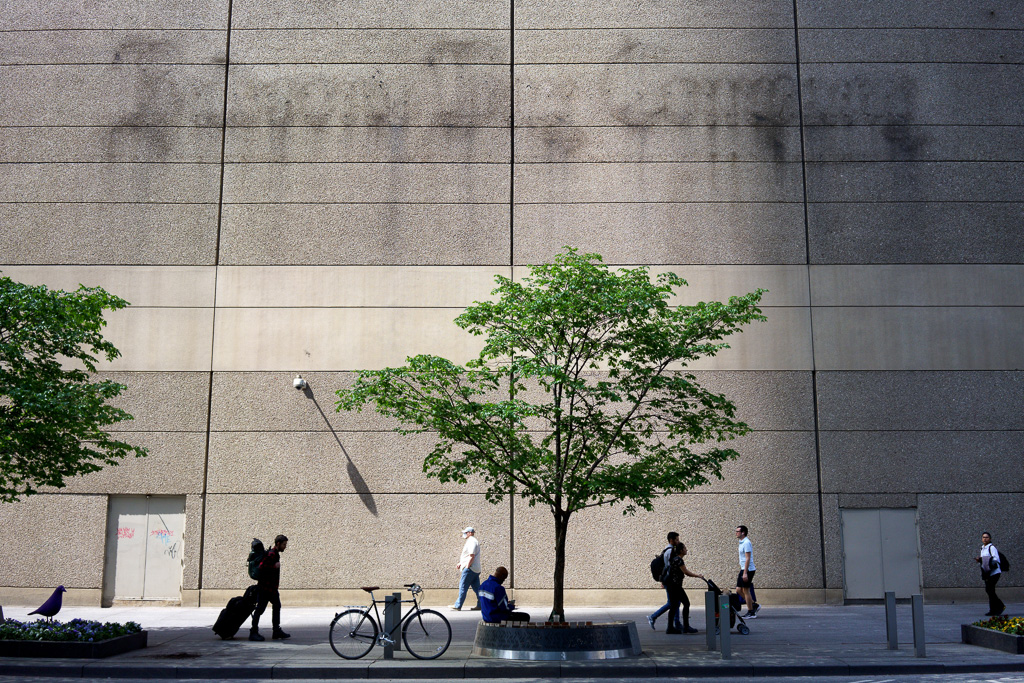 Shadowy outlines of the words "Hudson Bay Company" remain on the wall of the former Bloor/Yonge retail location after the sign has been dismantled. People pass on the sidewalk underneath.