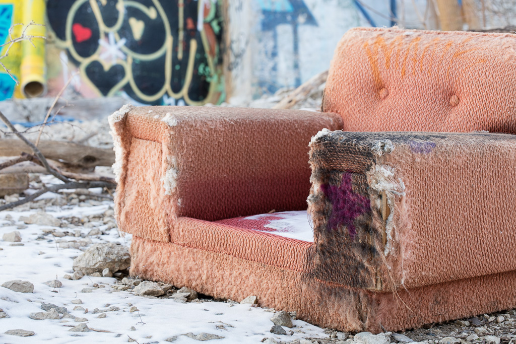 A ragged comfy chair sits outdoors on gravel with a light dusting of snow. In the background is a wall covered in graffiti.