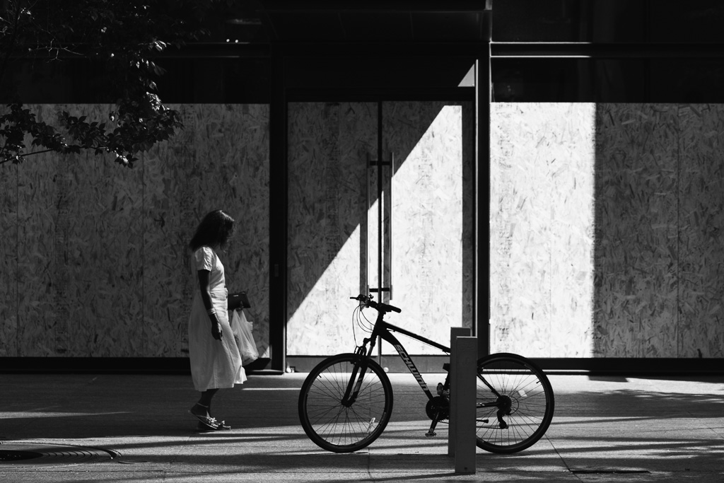 Woman passes plywood-covered shop windows on Bloor Street. A diagonal shadow terminates at her feet.