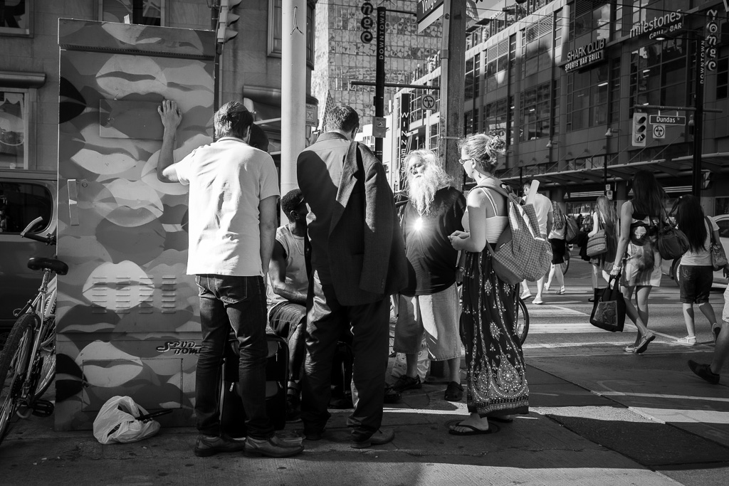 A street preacher with white hair and beard speaks to a gathering of youth on a street corner. Sunlight glints off a cross he wears and it looks as if his chest is on fire.