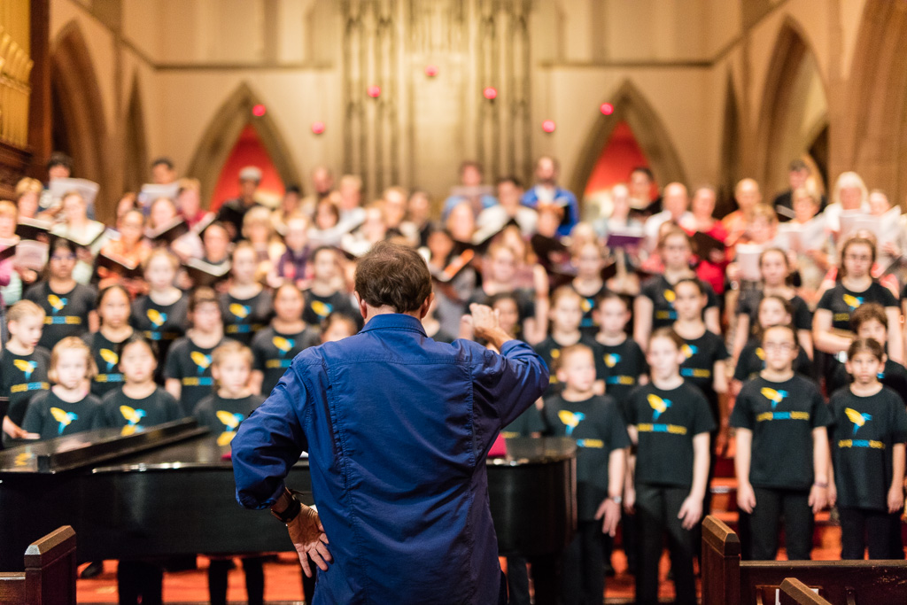 A choral conductor in casual dress stands before a grand piano and choir, right arm raised, left hand resting on his hip