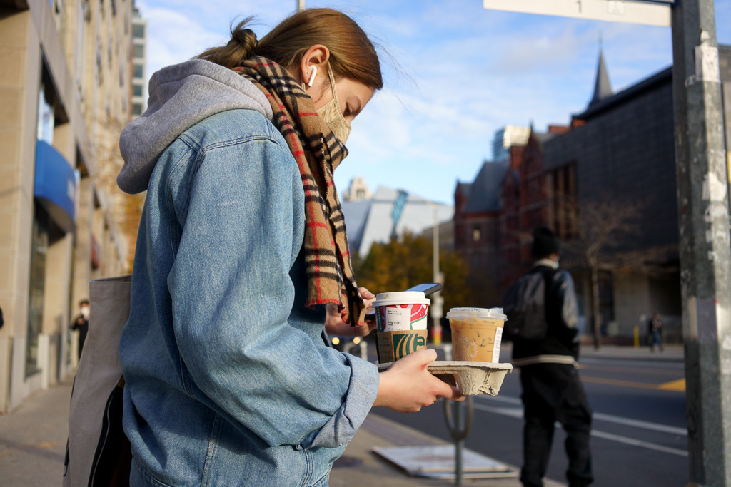 A woman texts with her left hand while holding a tray of Starbucks drinks in her right hand.