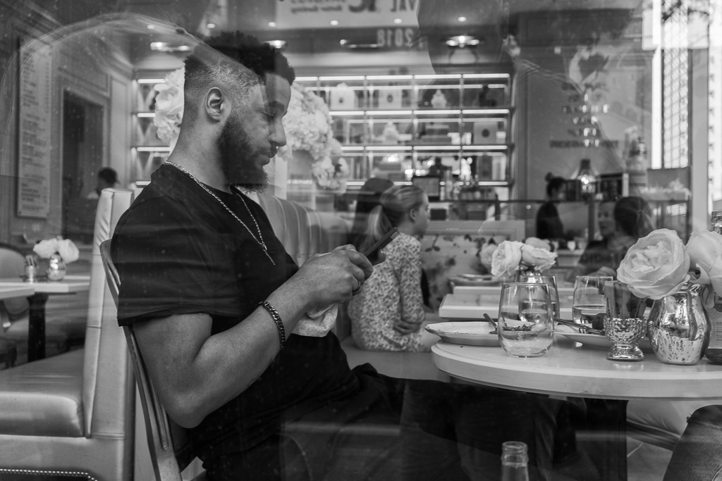 Black & white photo through a window of a man sitting at a restaurant table and texting.