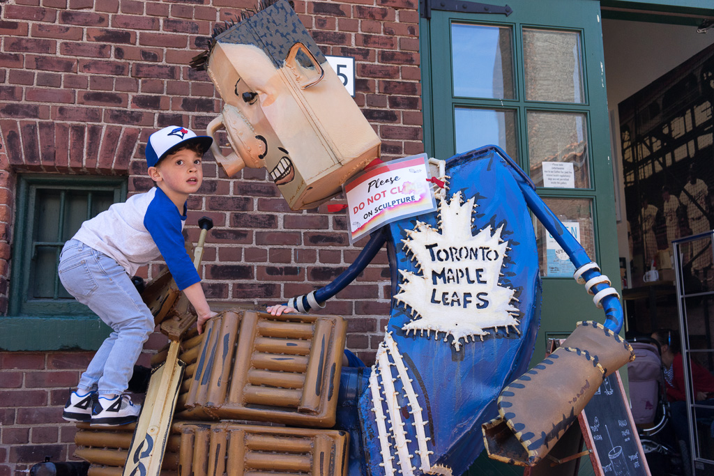 A child wearing a Toronto Blue Jays hat climbs on a sculpture of a Toronto Maple Leafs goalie with a sign that says: Please Do Not Climb On Sculpture