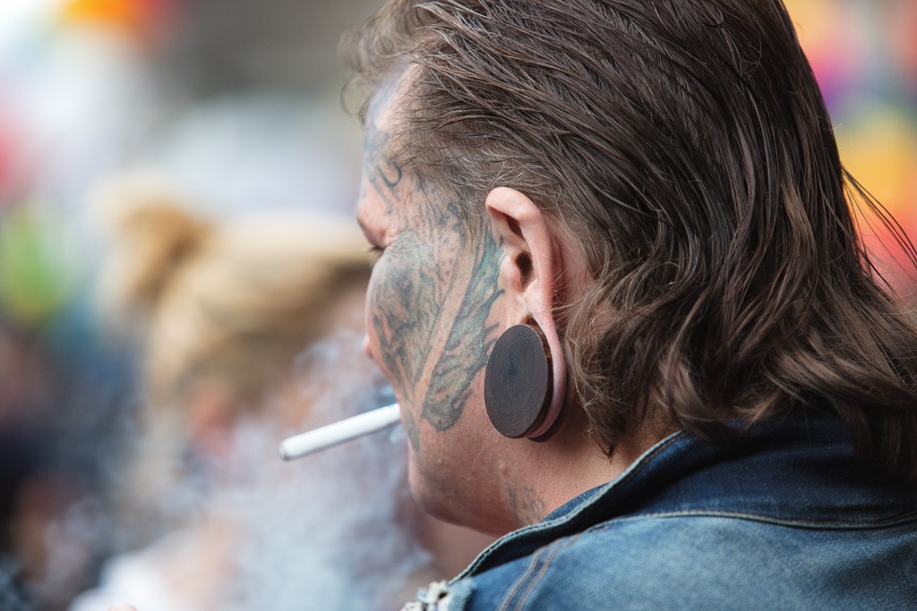 Candid street portrait from behind, focussing on ear lobes and facial tattoos. The subject has hair slicked back and smokes a cigarette. The smoke obscures an already blurred background.