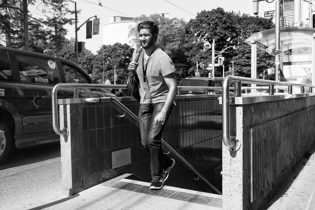 A young man in jeans and T-shirt and wearing a lanyard with ID card emerges onto the sidewalk from an underground subway entrance.