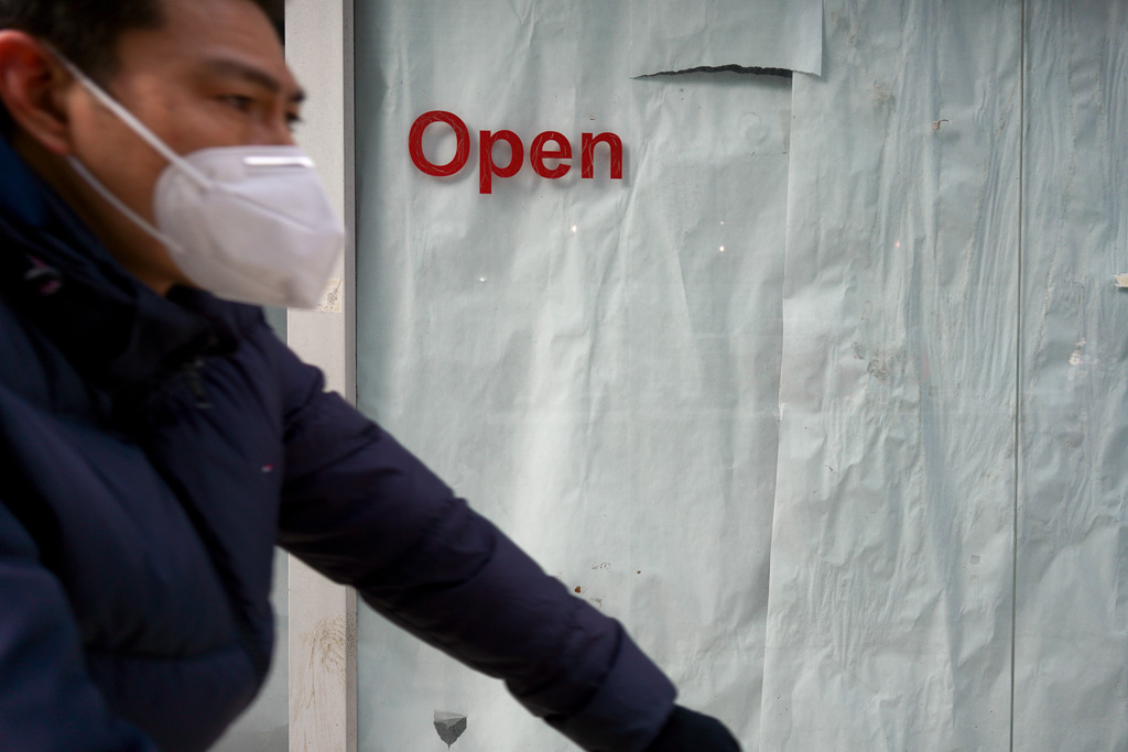 A man wearing a mask passes a store window that has been papered over but has the word "open" in red letters still stuck to the glass.