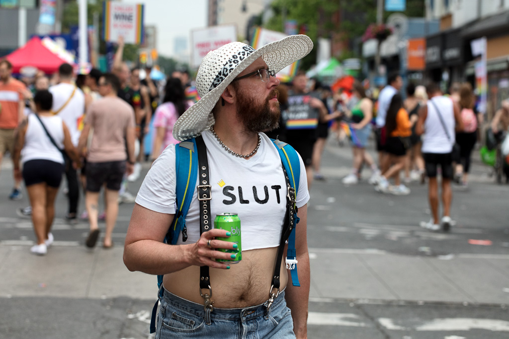 A person wearing a sun hat and a T-shirt with the word "Slut" on it walks down the middle of Toronto's Church Street holding a soft drink.