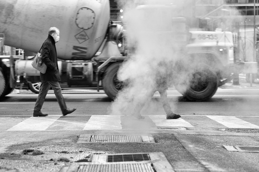 A black and white photo of a man walking past a steam vent while a cement truck passes in the background.