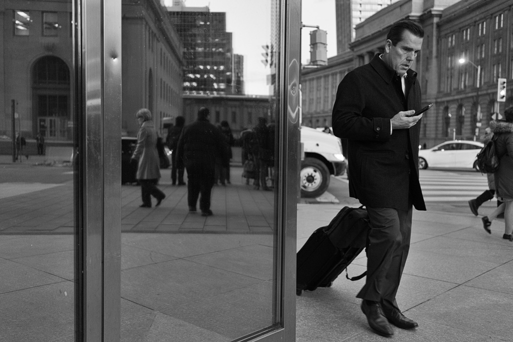 Black and white photograph of a man in business attire pulling a roller bag behind him with one hand while glancing at a smart phone in the other hand.