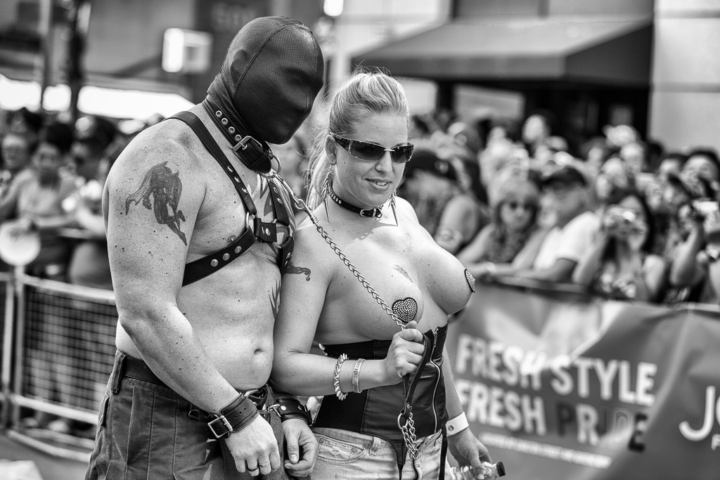 A topless woman leads a leather-clad man by leash and collar.