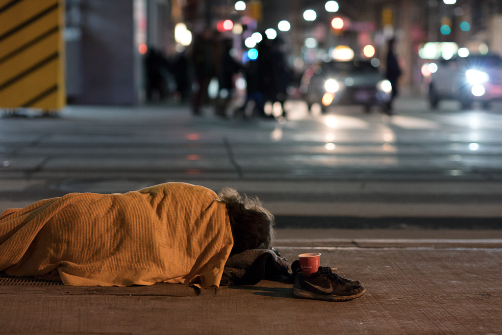 Night photograph of a homeless person sleeping underneath an orange blanket on the sidewalk at the intersection of Queen and Bay in Toronto.