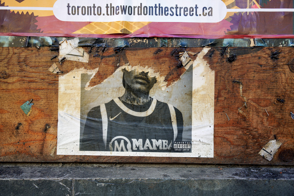 Photograph of posters on plywood. The bottom of a poster advertising Toronto's Word On The Street festival. Below it is a poster of a young man, tattoos on his neck, torn so that the top half of his head is missing. In the lower right corner of this poster is a parental advisory: explicit content.